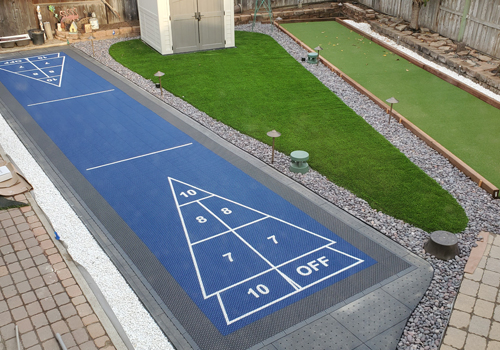 Blue shuffleboard court and turf bocce court in small backyard with shed and artificial turf
